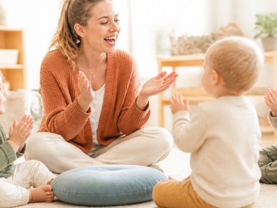 Erzieherin und drei Kleinkinder klatschen gemeinsam im Morgenkreis einer Kinderkrippe.